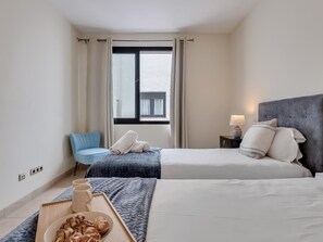 A hotel room with two beds, a small table with a tray of pastries, a chair, and a window with curtains. - Apartment in the heart of Puerto Banús - 4 guests (Marbella)