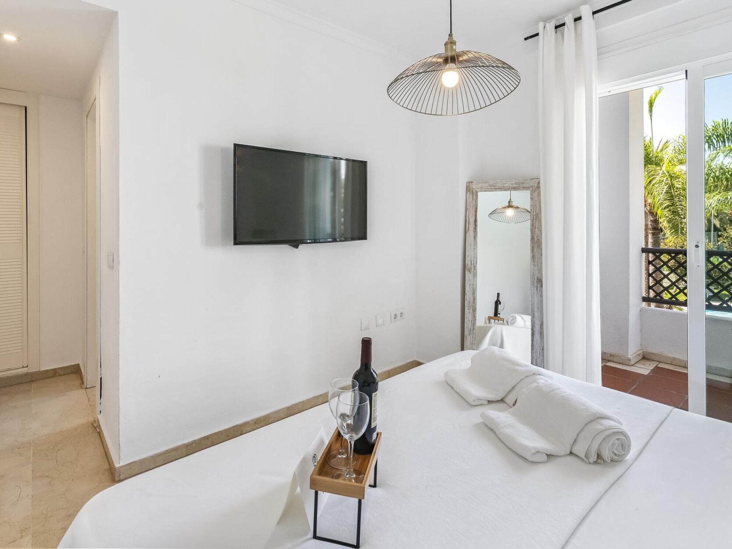 Habitación de hotel con cama, televisor montado en la pared, una botella de vino sobre una bandeja y vistas al exterior.