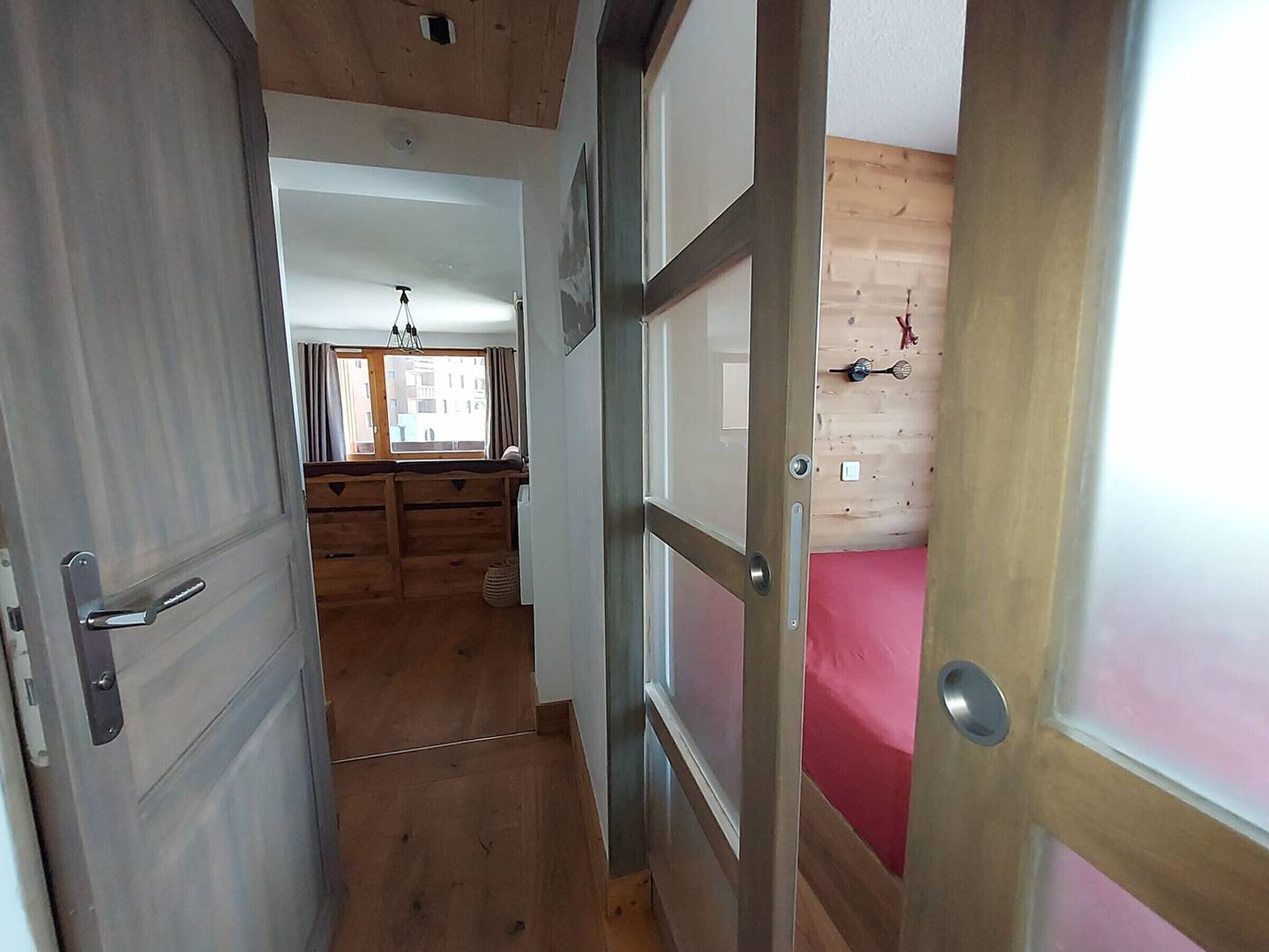 Ein schmaler Korridor mit Holzdielen führt zu einem Zimmer mit Bett und Fenster.
