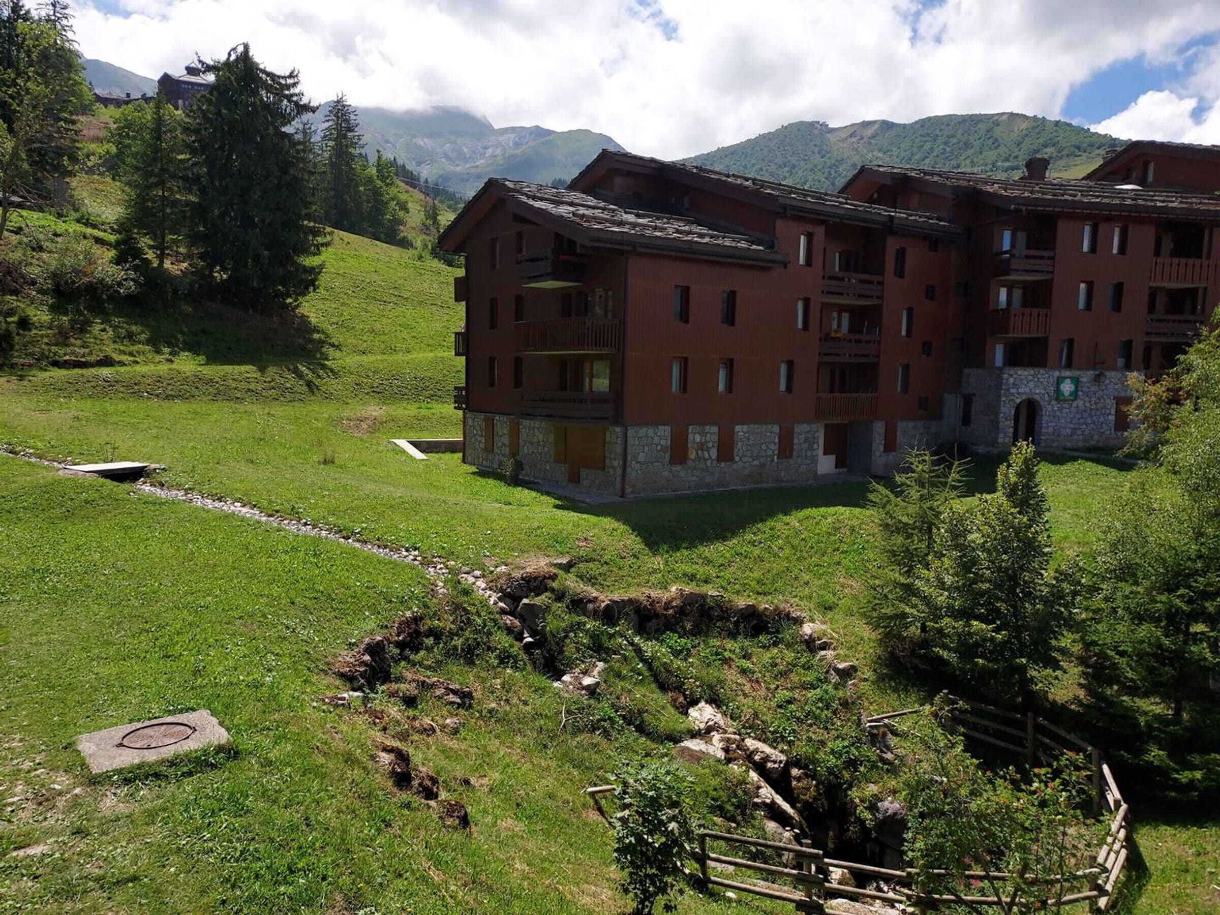 A multi-story building with balconies, surrounded by lush greenery and mountains in the background.