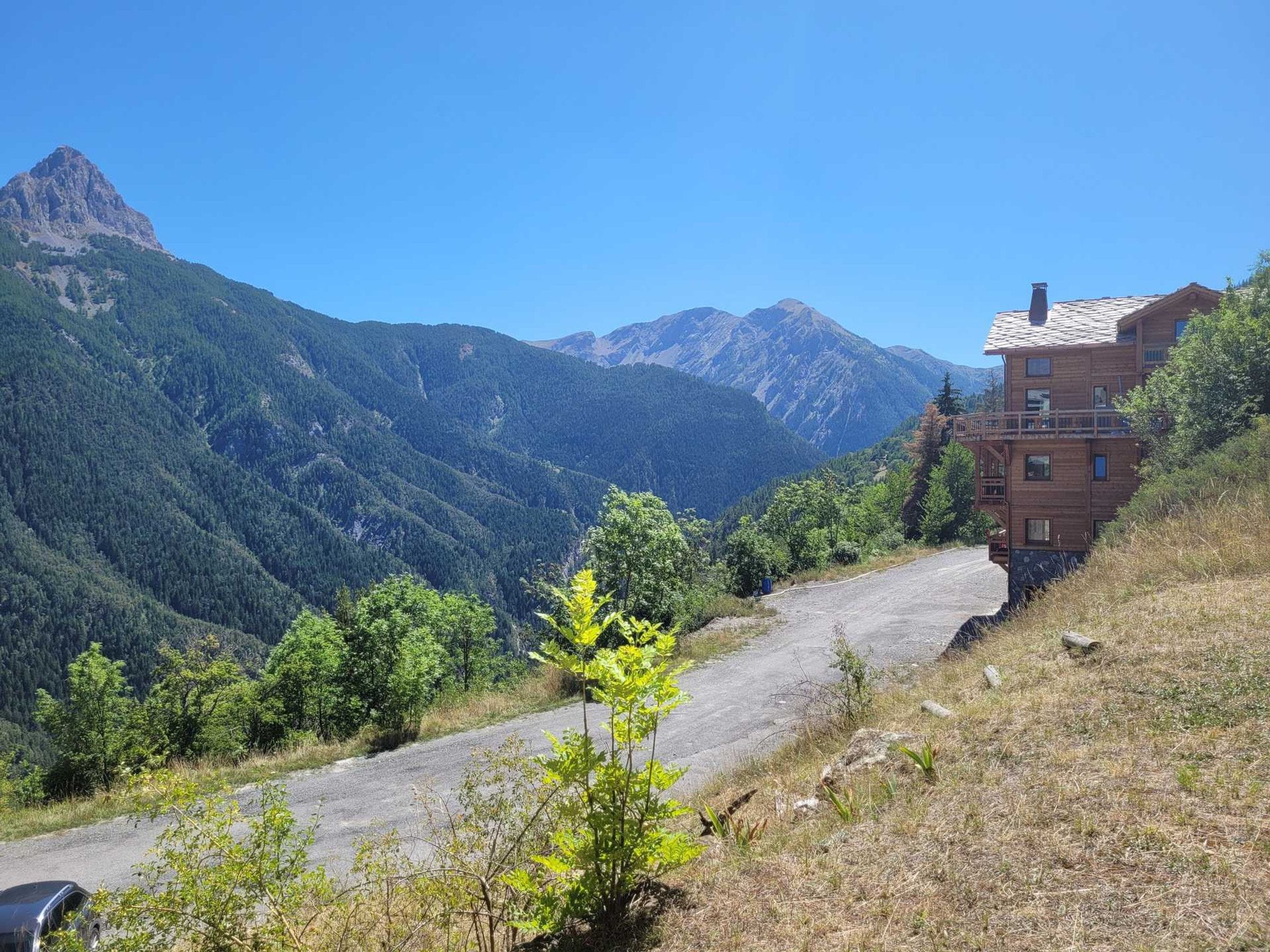 A mountain road leading to a wooden building with a balcony, surrounded by forested hills and a clear sky.