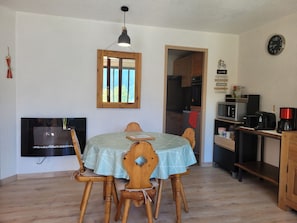 Ein Essbereich mit Tisch für vier Personen, Holzstühle, einer hängenden Pendelleuchte, einem an der Wand befestigten Fernseher und einer durchgehenden Tür erkennbaren Küche.