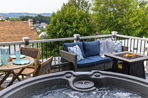 A hot tub on a balcony with a wicker sofa and chairs, a table with glasses, and a view of trees and buildings. - Premium Mt. Washington Retreat: VIP Views, Hot Tub, Garage (Pittsburgh)