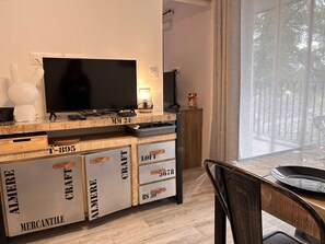 Ein modernes Wohnzimmer mit einem Flachbildfernseher, Holzschränken mit beschrifteten Fächern und einem Essbereich mit Tisch und Stühlen.