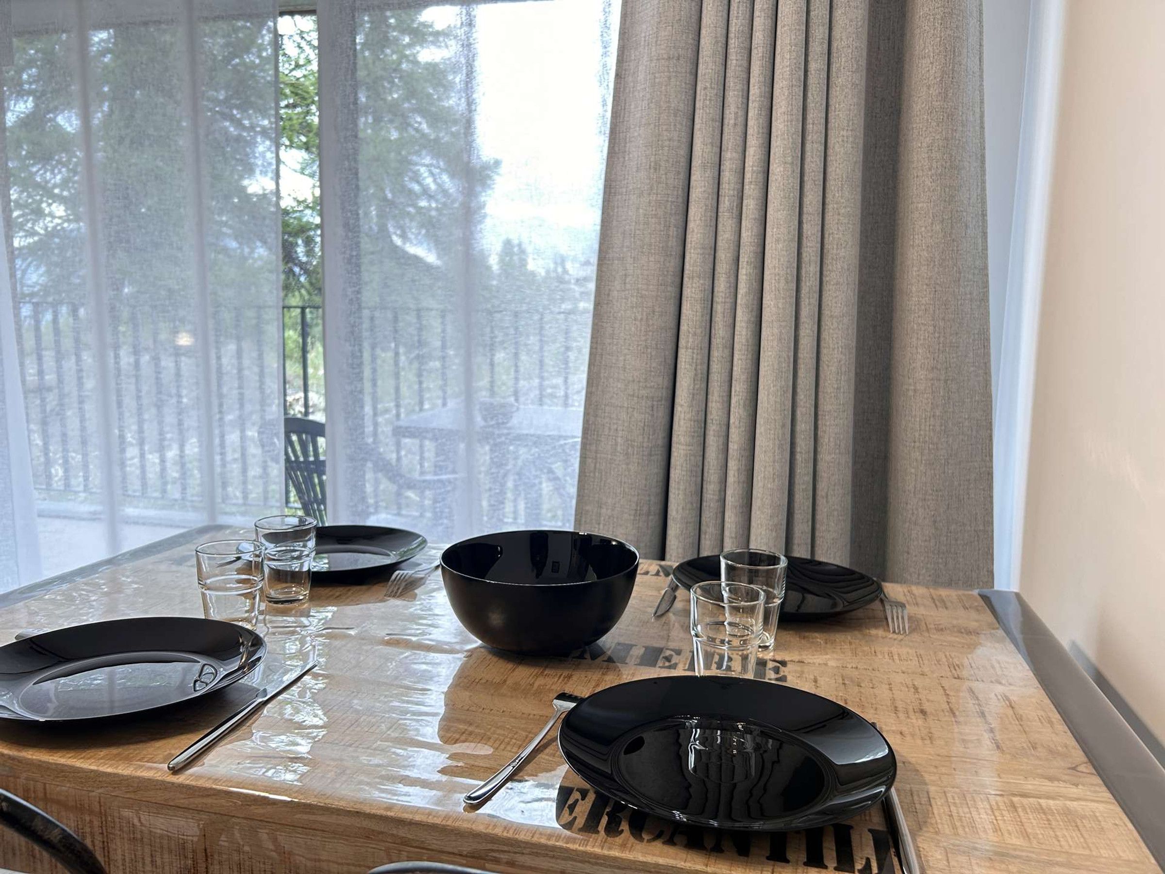 A dining table set with black plates, bowls, and cutlery, with a view of greenery outside.