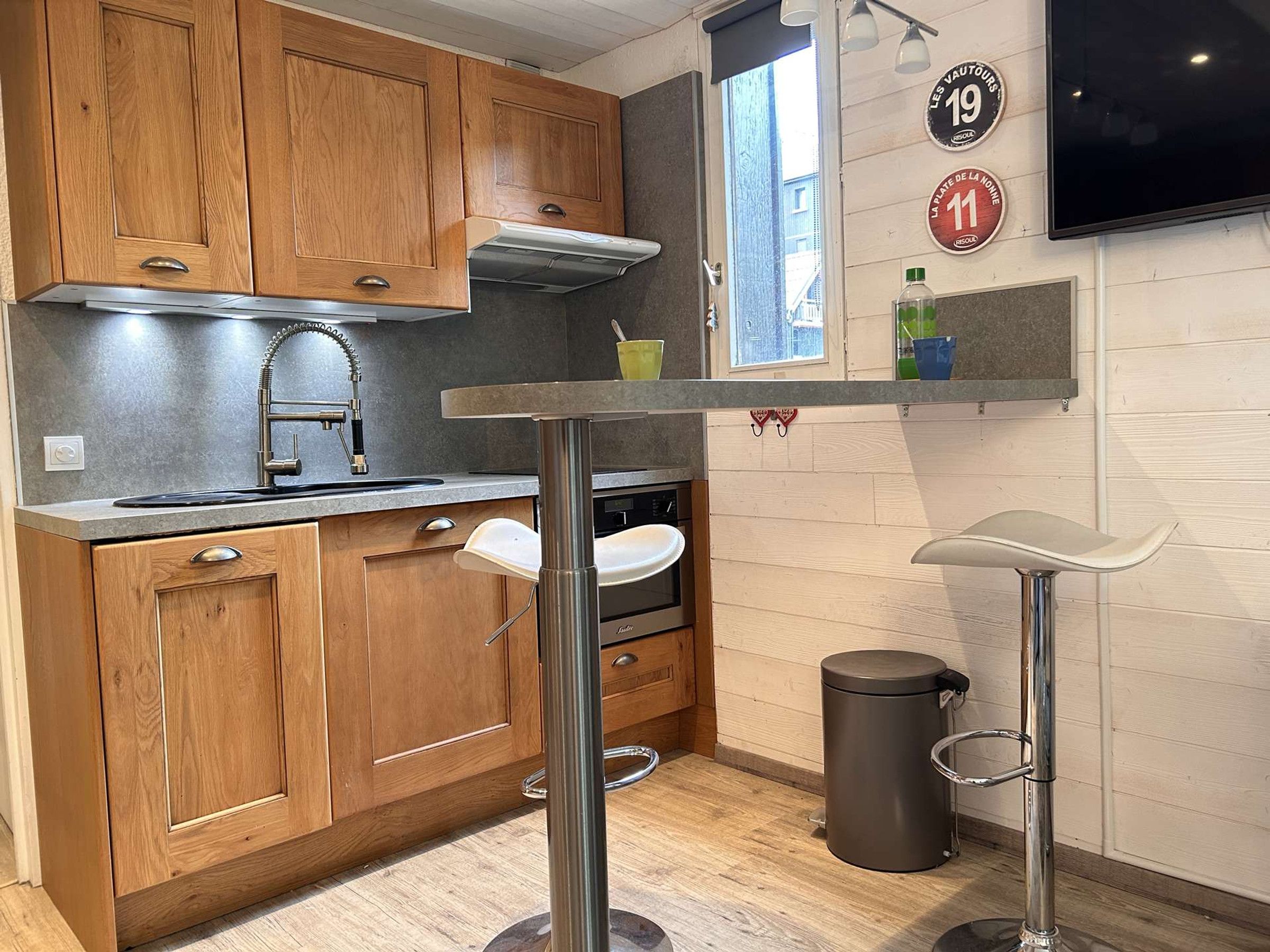 A compact kitchen with wooden cabinets, a stainless steel sink, a built-in oven, a small bar stool, and a trash bin.