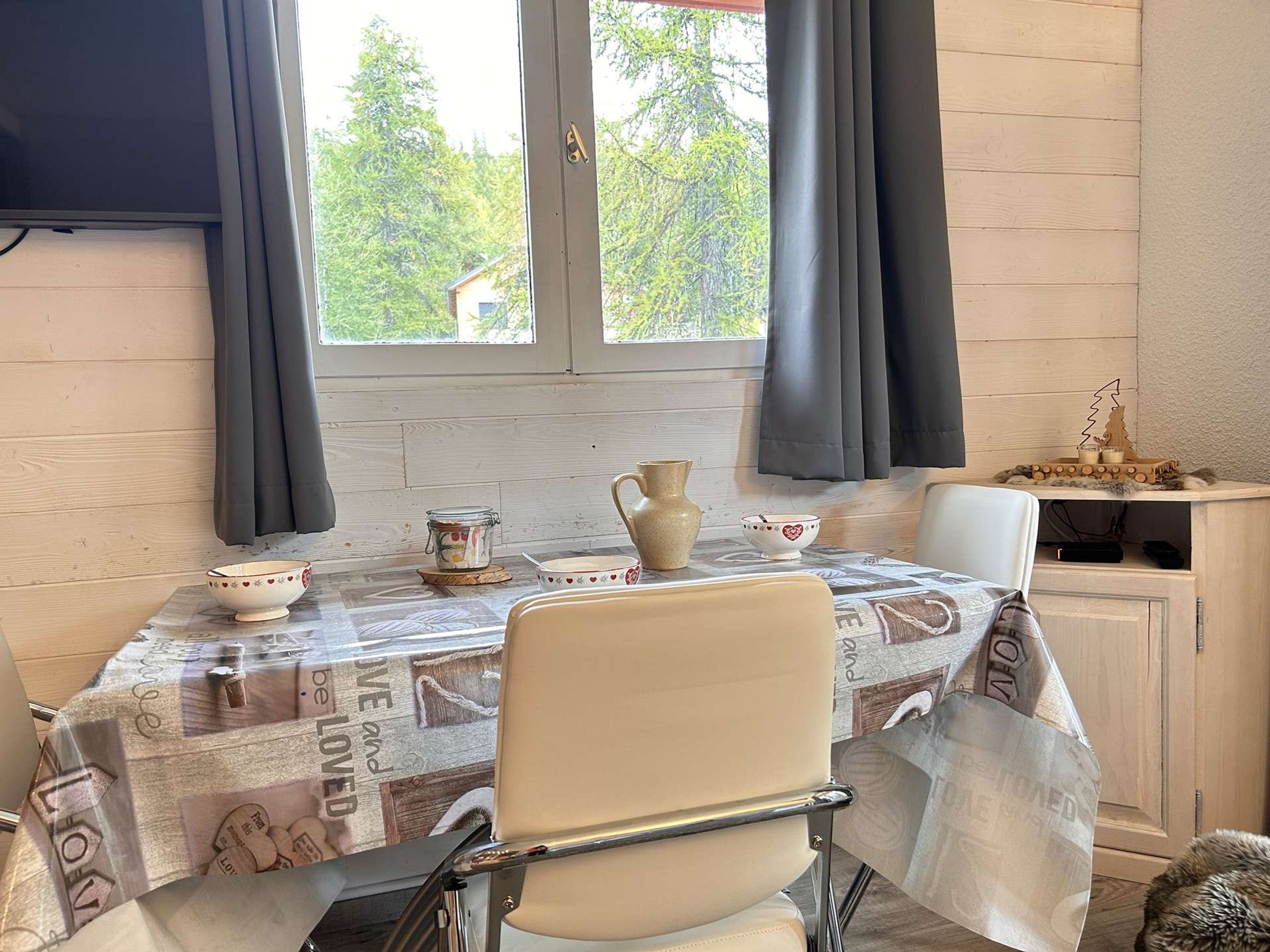 A dining area with a table set for two, a chair, a cabinet with a decorative item, and a window with curtains.