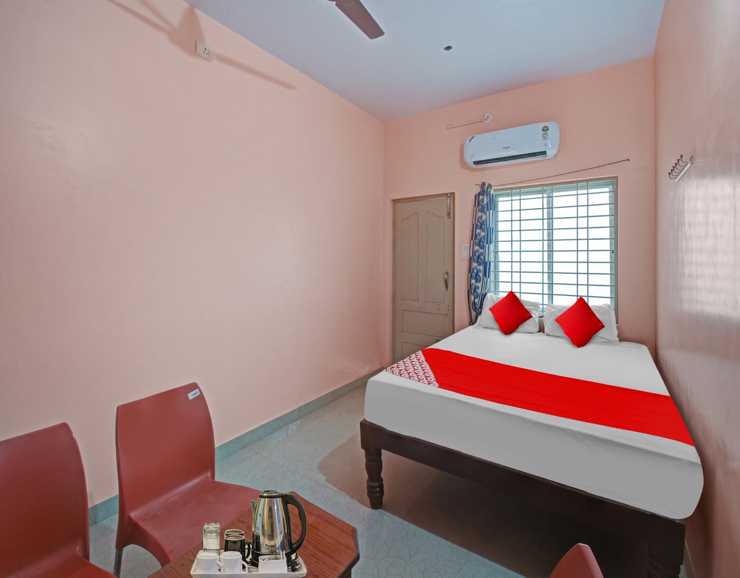 A hotel room with a bed, two chairs, a table with a kettle, and an air conditioning unit.