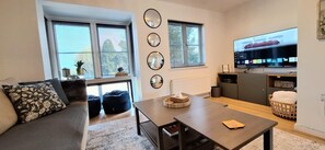Un salon moderne avec un canapé, une table basse et un téléviseur. Il y a trois miroirs au mur et une fenêtre avec des stores.