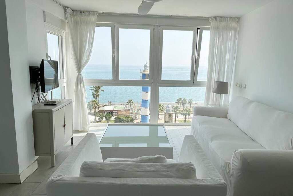 A room with a white sofa, a small table, and a TV on a cabinet. A large window offers a view of the sea and a lighthouse.
