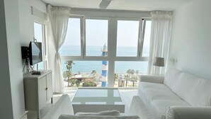 A room with a white sofa, a small table, and a TV on a cabinet. A large window offers a view of the sea and a lighthouse.