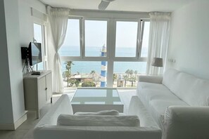 A room with a white sofa, a small table, and a TV on a cabinet. A large window offers a view of the sea and a lighthouse.