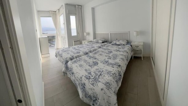 A bedroom with a bed, bedside tables, a ceiling fan, and a view of the sea.