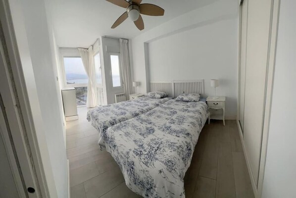 A bedroom with a bed, bedside tables, a ceiling fan, and a view of the sea.