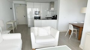 A modern, minimalist living room with a white sofa, a glass coffee table, and a dining area with a wooden table and chairs.