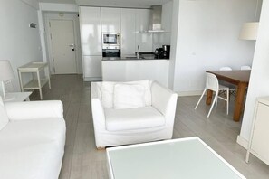 A modern, minimalist living room with a white sofa, a glass coffee table, and a dining area with a wooden table and chairs.
