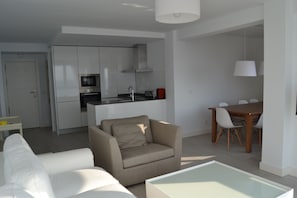 A modern living space with a sofa, a glass coffee table, a dining table with chairs, a kitchen area with cabinets and appliances, and a ceiling light fixture. - Piso del Mar. Stunning views just 50 meters from the beach (Torre del Mar)