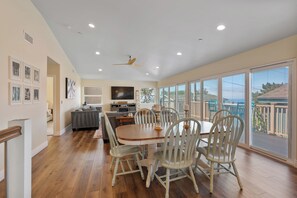 A spacious dining area with a wooden table and white chairs, a ceiling fan, and large windows offering a view of the outdoors. - The Otter House on Happy Hill! Ocean Views & Short Walk to Moonstone Beach! (CAMBRIA)