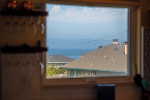 A window with a view of a residential area, a clear sky, and a distant body of water. - The Otter House on Happy Hill! Ocean Views & Short Walk to Moonstone Beach! (CAMBRIA)