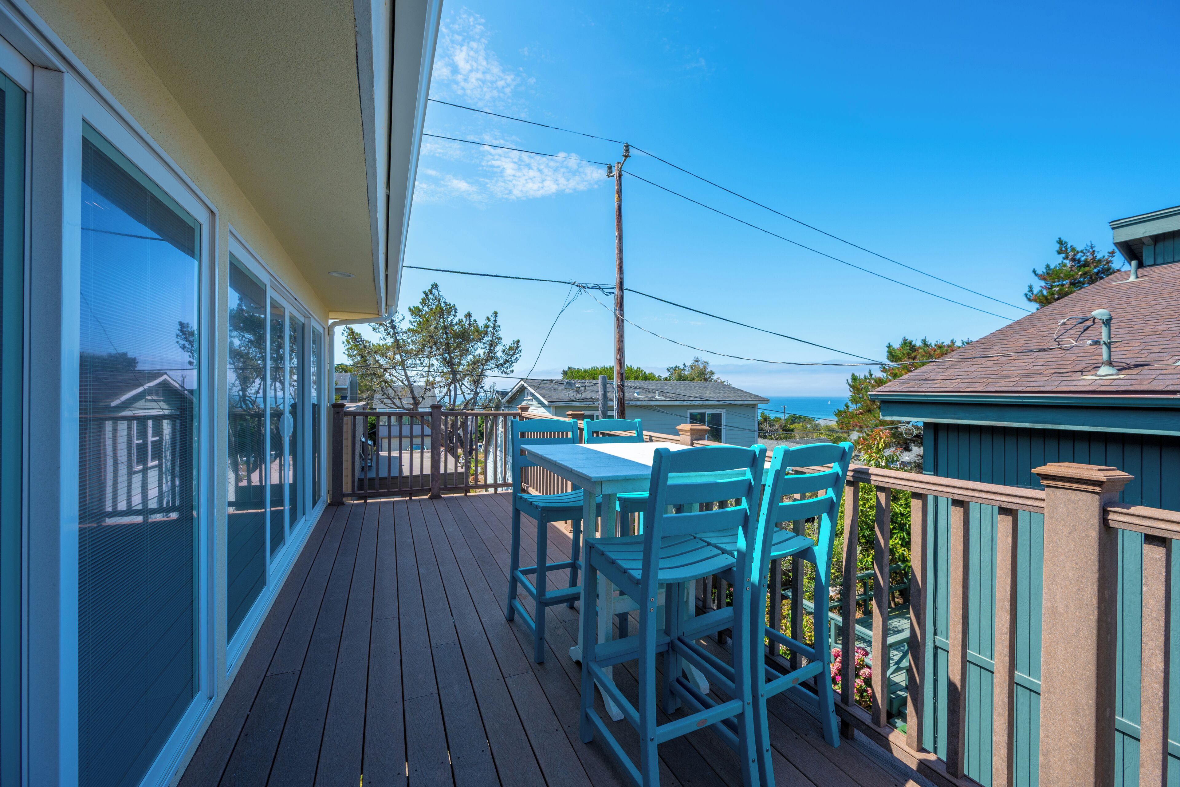 Eine Holzterrasse mit Tisch und Stühlen, mit Blick auf einen Wohnbereich und das Meer.
