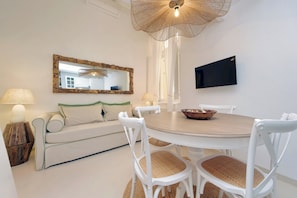 Un coin repas avec une table ronde, des chaises blanches, une petite table d’appoint en bois, un miroir et un téléviseur fixé au mur.