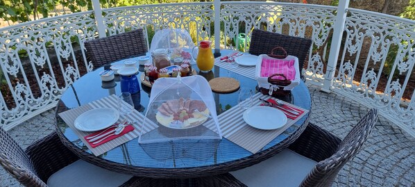 A round table set for a meal with wicker chairs, a glass table, and a white metal fence in the background.