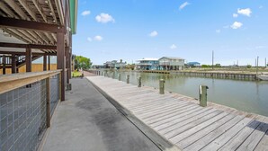 A wooden pier leading to a waterfront property with a covered area and a view of houses across the water. - SeaGlass Smiles | Canal-Front, Private Dock & Dog-Friendly (Rockport)