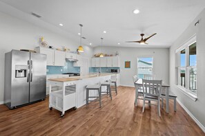 A modern kitchen with white cabinetry, a stainless steel refrigerator, a wooden island with bar stools, and a view of the outside through a large window. - Fall Special! Buy 2 nights, get your 3rd night Free! At Seaglass Smiles (Rockport)