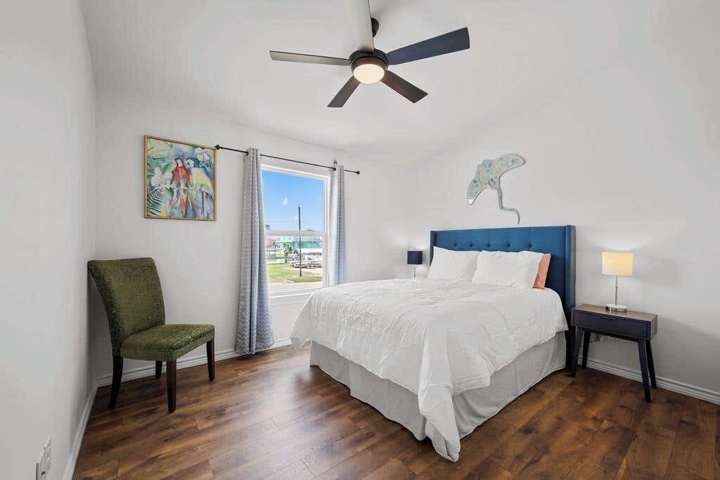 Een slaapkamer met een bed, een stoel, een nachtkastje, een plafondventilator, een raam met gordijnen en een schilderij aan de muur.