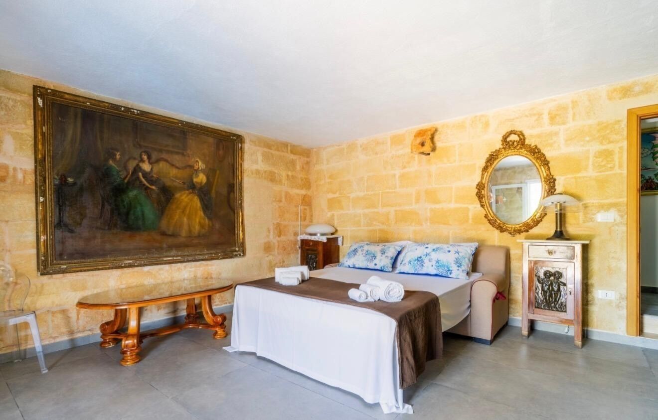 A hotel room with a large painting, a bed with a blue floral bedspread, a bench, and a wooden nightstand with a lamp.