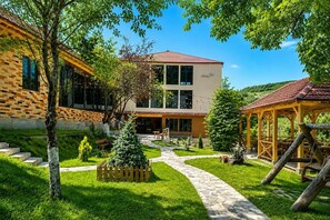 A two-story building with a brick facade and a tiled roof, surrounded by a well-maintained garden with a pathway leading to a wooden gazebo.