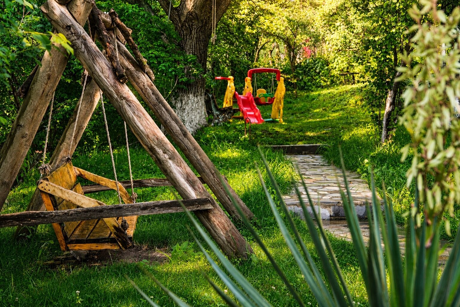 A rustic wooden swing set in a lush green garden with a stone pathway.