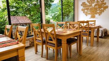 Un espace repas avec des tables et des chaises en bois, de grandes fenêtres donnant sur un paysage verdoyant et un schéma représentant un arbre, fixé au mur.