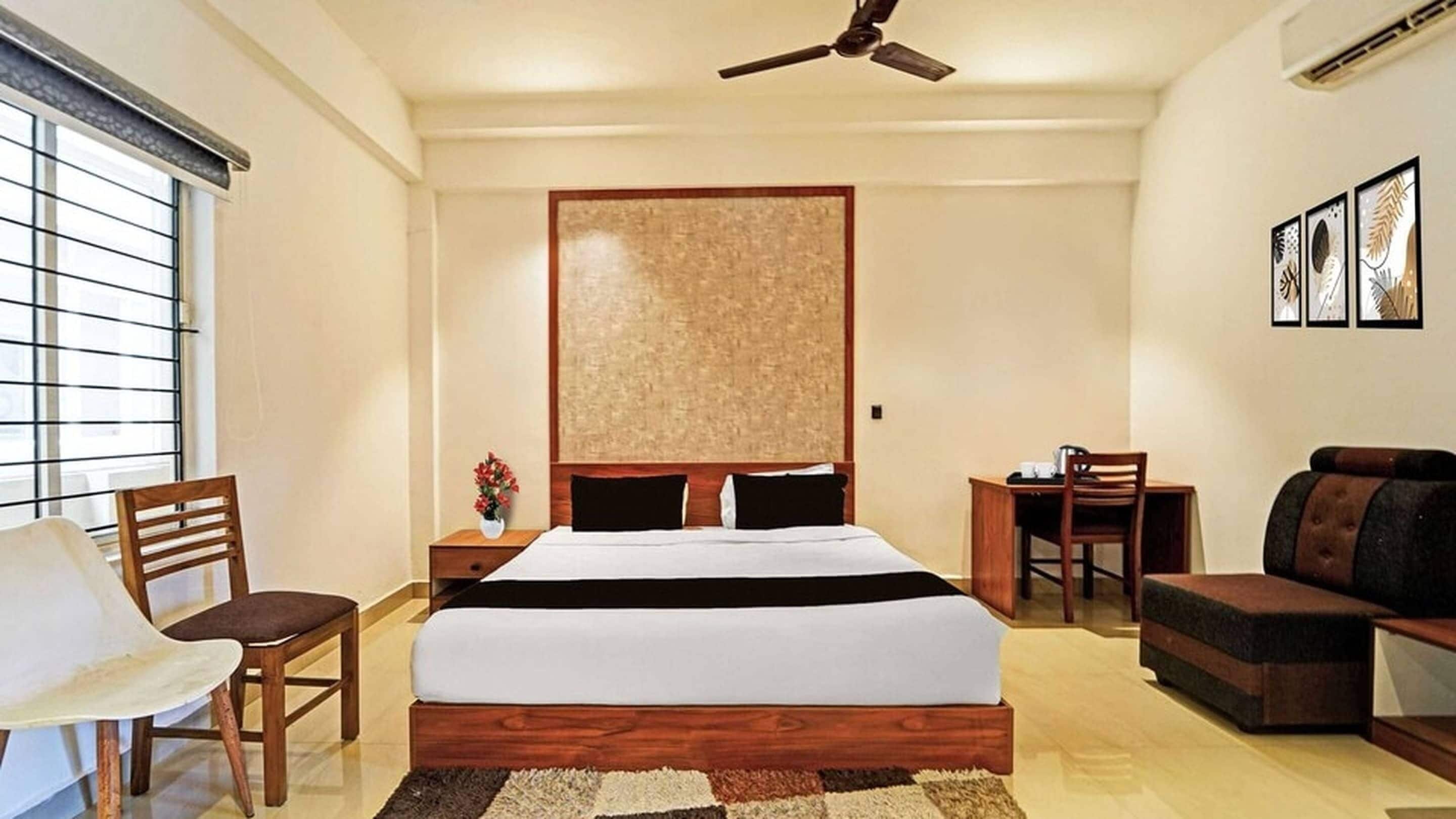 A hotel room with a bed, a desk, a chair, a sofa, a ceiling fan, and a window with blinds.