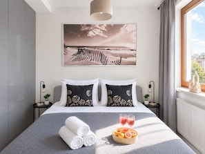 A hotel room with a bed, pillows, a bedside table with a lamp, a window with curtains, and a wall-mounted picture. - Lumina 2BR Apart with Parking AC Balcony (Warszawa)