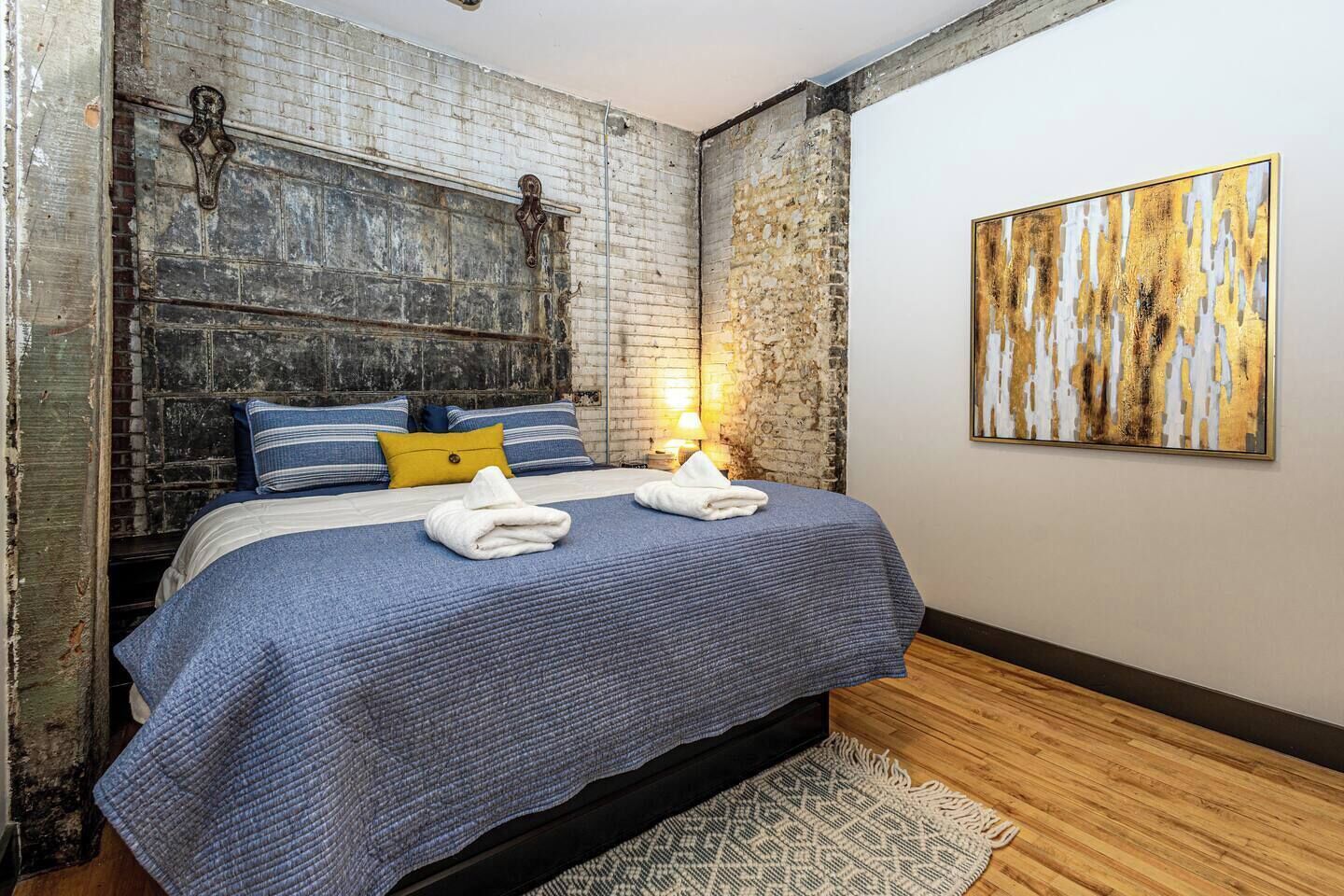 Ein Schlafzimmer mit Backsteinwand, einem Bett mit blauer Bettwäsche, einem gelben Kopfkissen und einem Bild an der Wand.