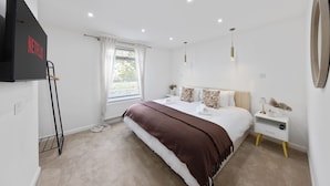A modern bedroom with a bed, bedside tables, a TV, and a window with curtains. - Stylish Home Near Chelsfield Station Free Wifi (Orpington)