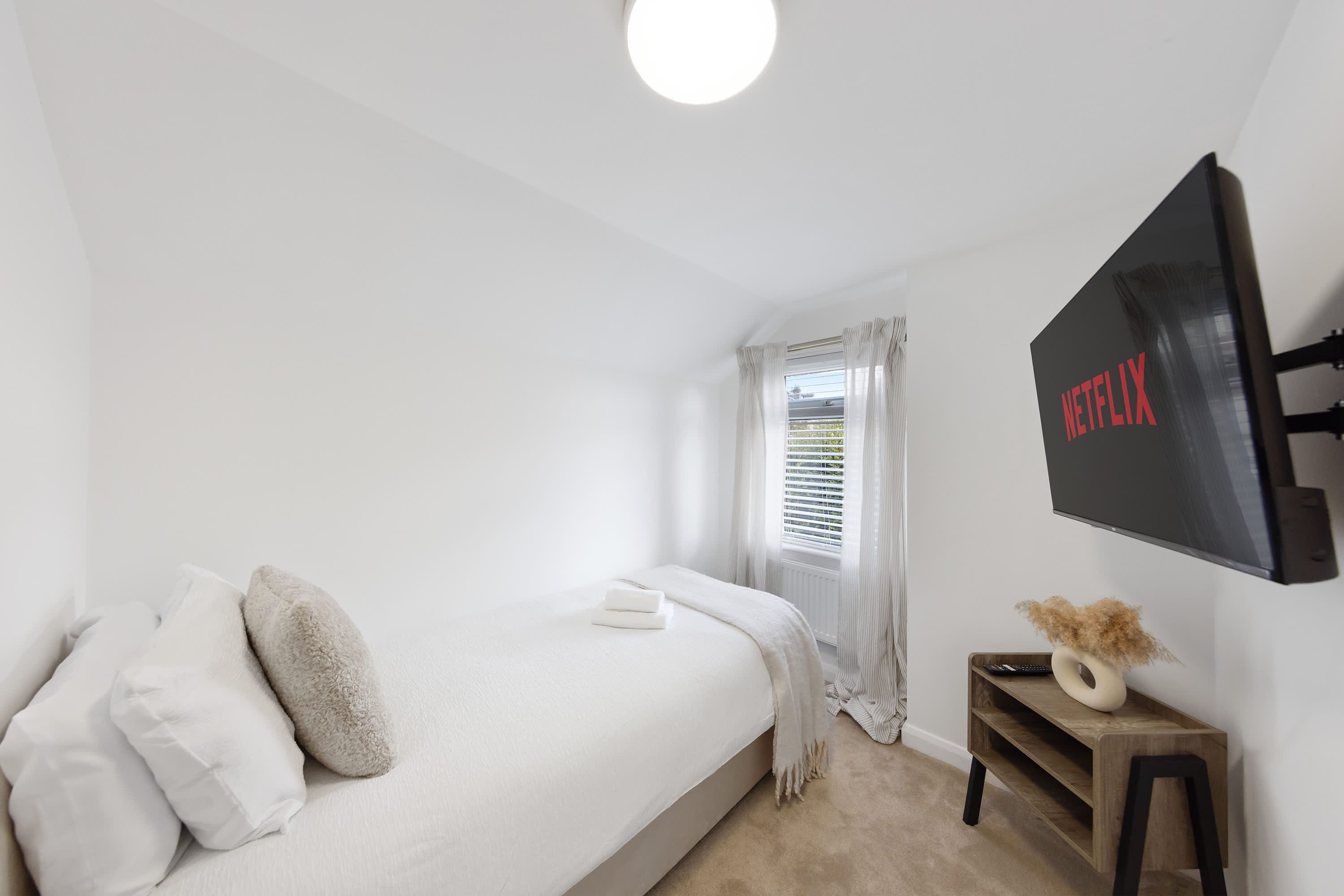 Ein Schlafzimmer mit einem Bett, einem an der Wand befestigten Fernseher, einem Fenster mit Vorhängen und einem Nachttisch mit einer Vase.