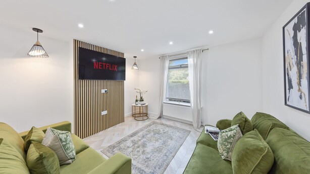 A modern living room with a green sofa set, a flat-screen TV mounted on a wooden panel, a small side table with a bottle and glasses, and a window with white curtains. - Stylish Home Near Chelsfield Station Free Wifi (Orpington)