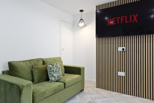 Un sofá verde con un cojín estampado, un letrero de Netflix fijado a la pared y un revestimiento de pared de listones de madera.