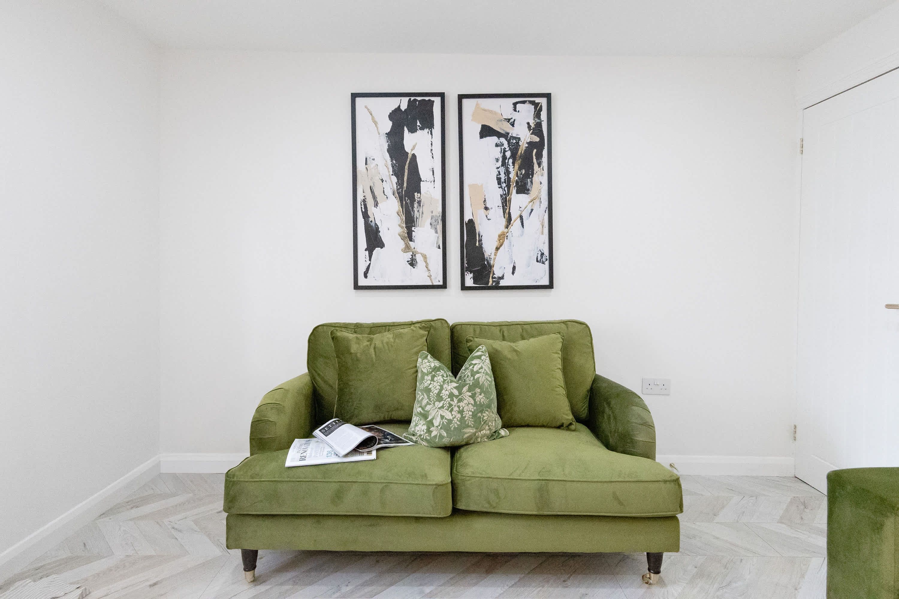 A green sofa with floral cushions, a magazine, and two abstract paintings on the wall.