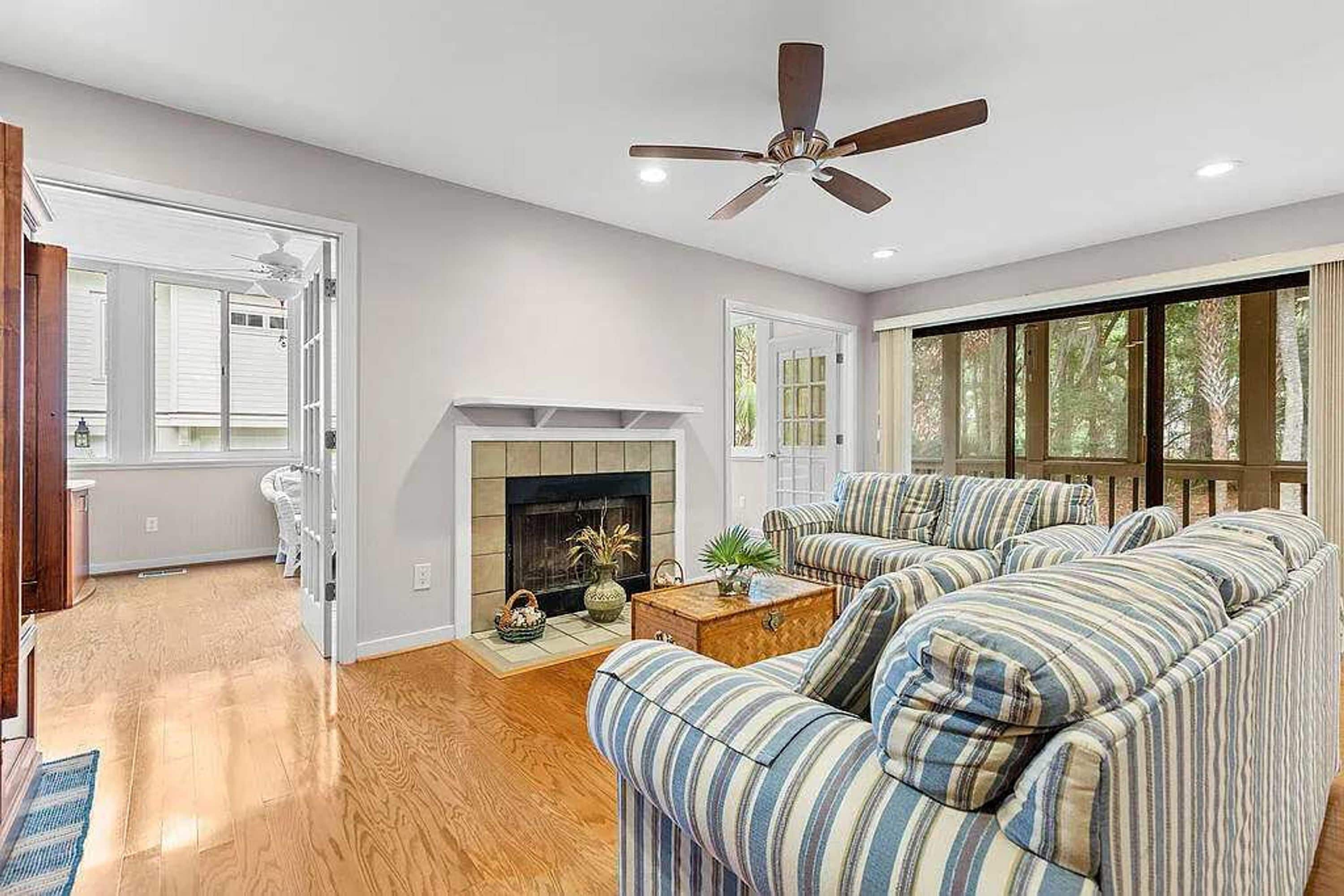 A living room with a fireplace, striped sofas, a ceiling fan, and large windows.