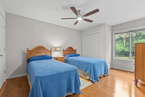 A bedroom with two beds, a ceiling fan, a window with a view of greenery, and a door. - Anchor Down - 2 Bed Beach Fun Dining Golf (Seabrook Island)