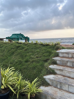 A coastal area with a grassy path leading to a beach, a covered seating area, and a concrete staircase.