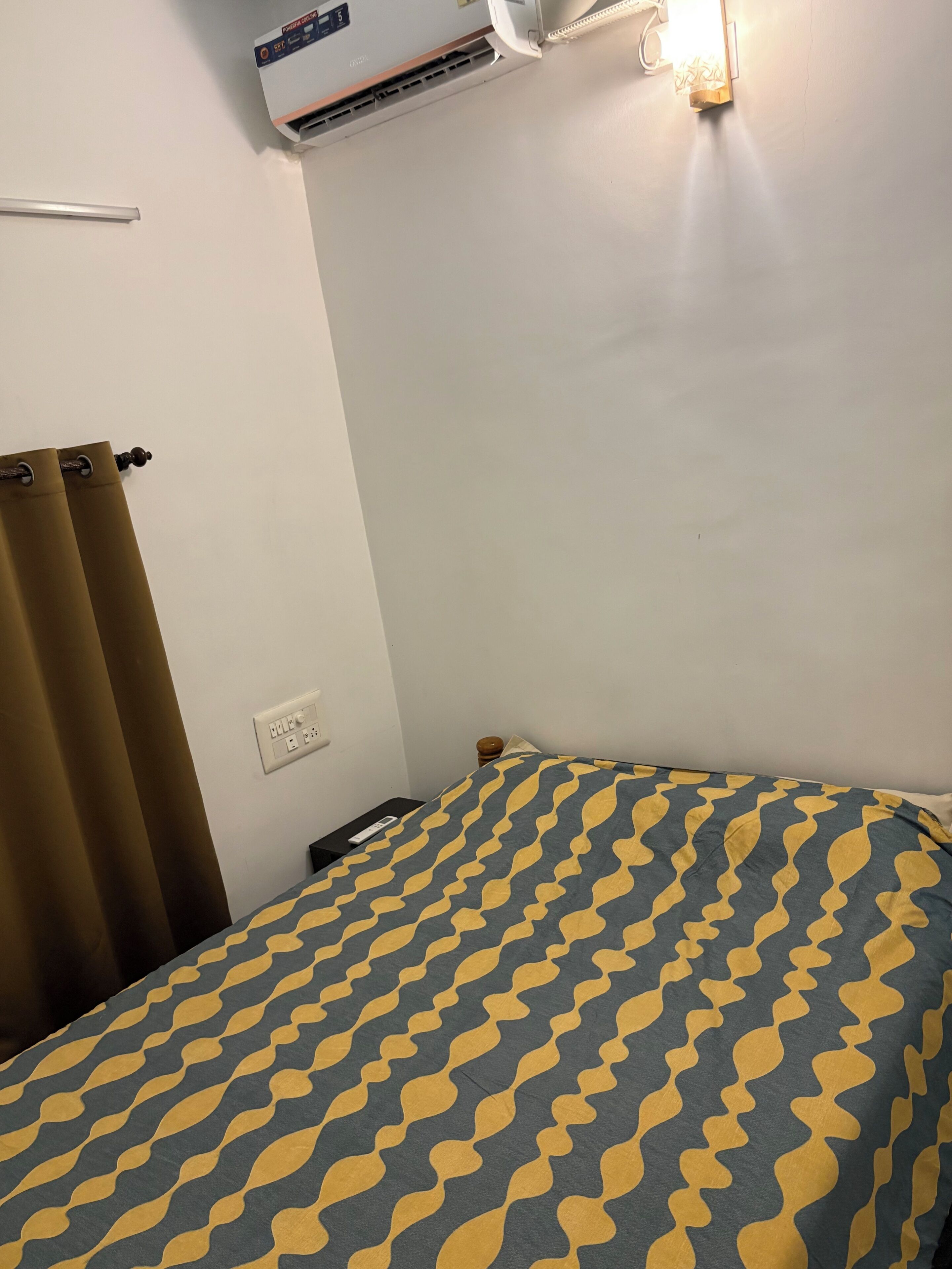 A hotel room with a bed, an air conditioning unit, a wall socket, and a remote control on the nightstand.