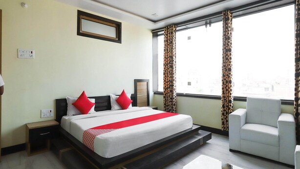 A hotel room with a bed, a chair, a nightstand, a bedside table, and a window with curtains.