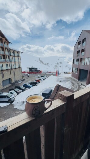 Un balcón con una taza de café, un estacionamiento nevado y edificios al fondo.
