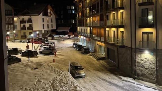 A snow-covered street with a building featuring balconies and a parking area.