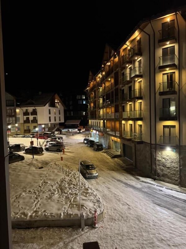 A snow-covered street with a building featuring balconies and a parking area.