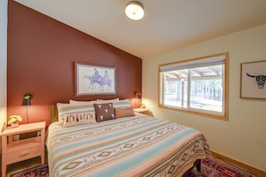 A bedroom with a large bed, a window with a view of trees, a mounted painting of horses, and a wall-mounted lamp.